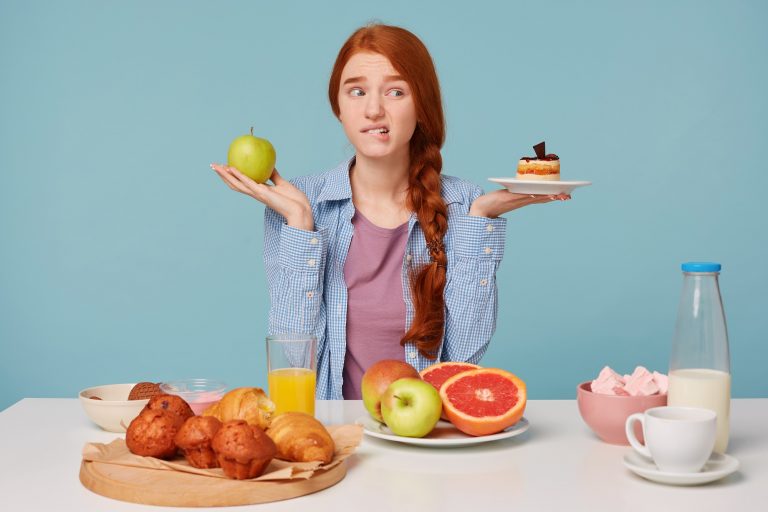 Hard choice. Portrait of beautiful woman trying to choose between healthy and unhealthy food while isolated on bluebackground. Woman holding cake and apple in hands